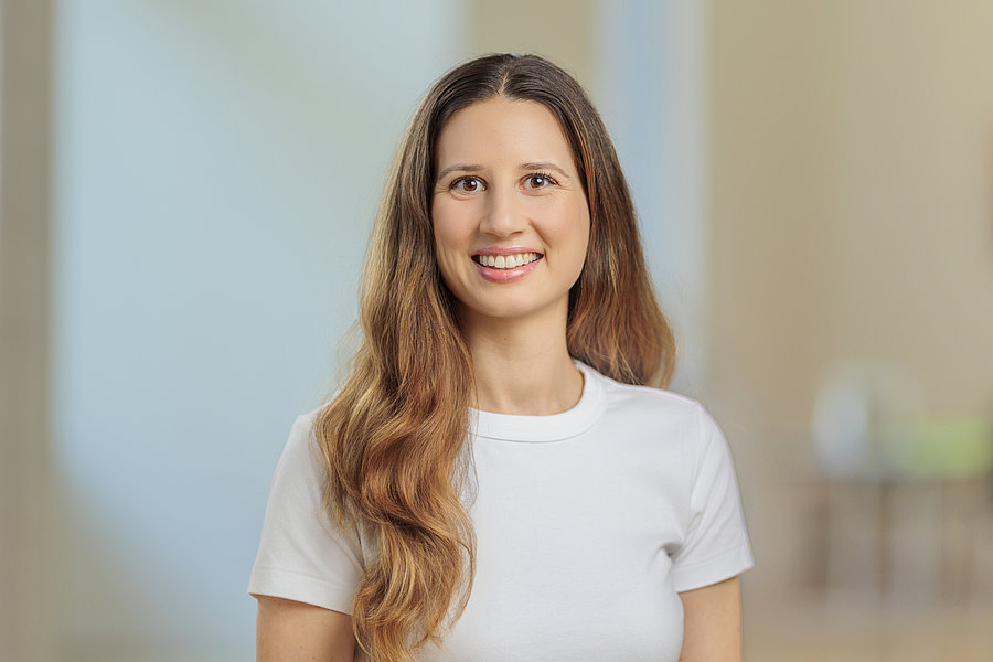 Frau mit langen, gewellten Haaren trägt ein weißes T-Shirt und steht vor einem unscharfen Hintergrund