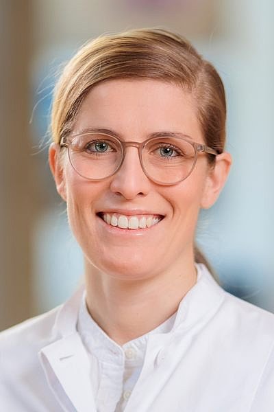 Person mit blondem, zurückgekämmtem Haar und weißem Hemd vor unscharfem Hintergrund
