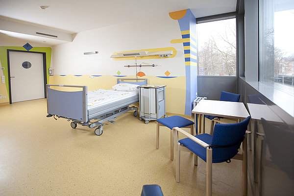 Krankenhauszimmer mit einem Bett, zwei blauen Stühlen und einem kleinen Tisch neben einem großen Fenster
