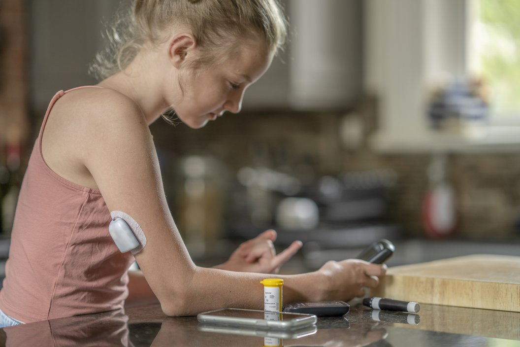 Ein Mädchen mit einem Sensor am Oberarm sitzt an einem Tisch und hält ein Smartphone in der Hand. Auf dem Tisch liegen eine Pillendose und ein weiteres Smartphone.