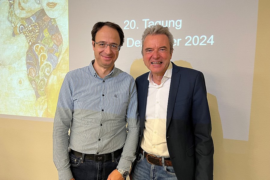 Dr. Korkolis und Dr. Berger stehen nebeneinander, beide in formeller Kleidung. Im Hintergrund ist ein Projektor mit dem Text '20. Tagung Dezember 2024' sichtbar.