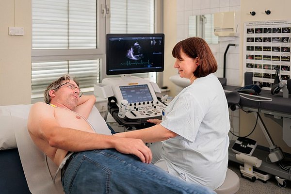 Ärztin führt Herzultraschall bei einem liegenden männlichen Patienten durch, Ultraschallbild auf Monitor sichtbar