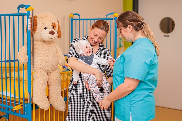Frau in blauem Pflegehemd hält die Hand eines Babys, das von einer anderen Frau in kariertem Kleid gehalten wird, neben einem Kinderbett mit großem Plüschhund