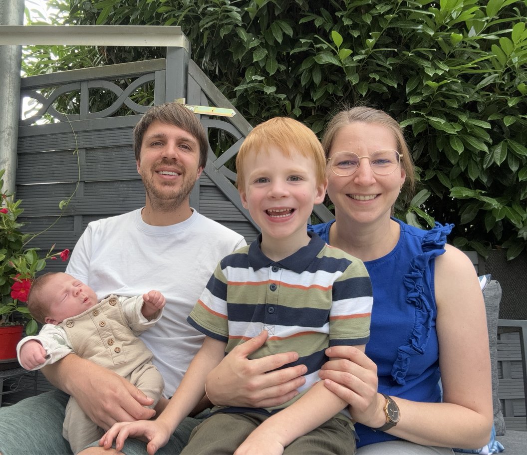 Eine Person hält ein Baby in einem weißen Anzug, ein Junge mit rotem Haar trägt ein gestreiftes T-Shirt, eine Frau in einem blauen Oberteil sitzt neben ihm. Im Hintergrund sind Pflanzen zu sehen.