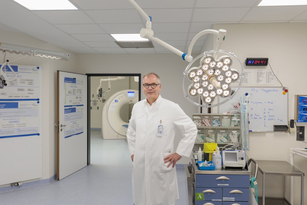 Ein Arzt in einem weißen Kittel steht mit den Händen in der Hüfte in einem Operationssaal. Im Hintergrund sind medizinische Geräte und ein CT-Scanner sichtbar.