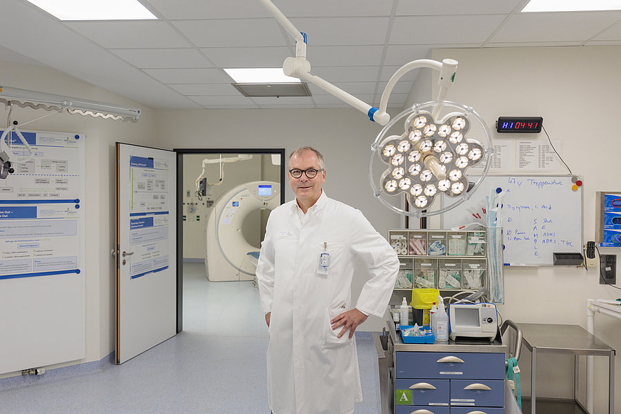 Ein Arzt in einem weißen Kittel steht mit den Händen in der Hüfte in einem Operationssaal. Im Hintergrund sind medizinische Geräte und ein CT-Scanner sichtbar.