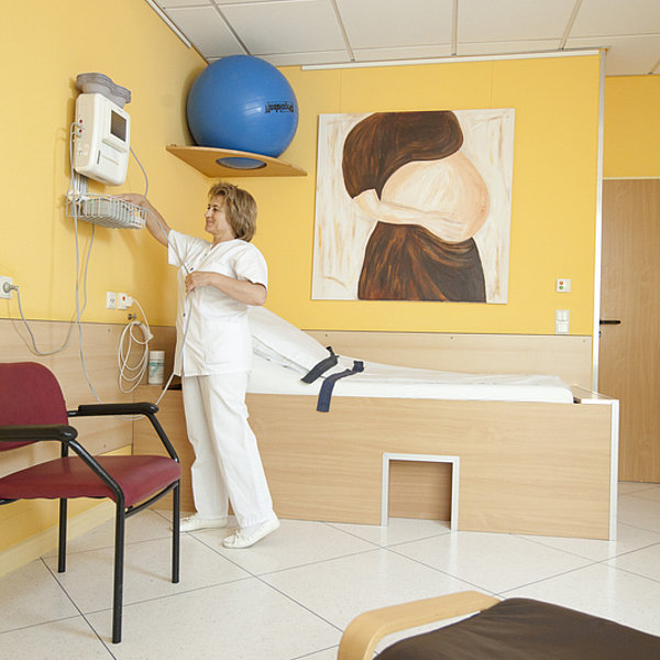 Pflegekraft in weißer Kleidung bedient medizinisches Gerät in gelbem Patientenzimmer mit Liege und Stuhl