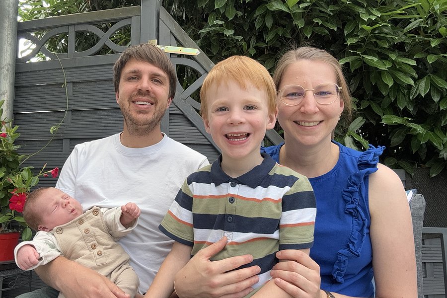 Eine Person hält ein Baby in einem weißen Anzug, ein Junge mit rotem Haar trägt ein gestreiftes T-Shirt, eine Frau in einem blauen Oberteil sitzt neben ihm. Im Hintergrund sind Pflanzen zu sehen.