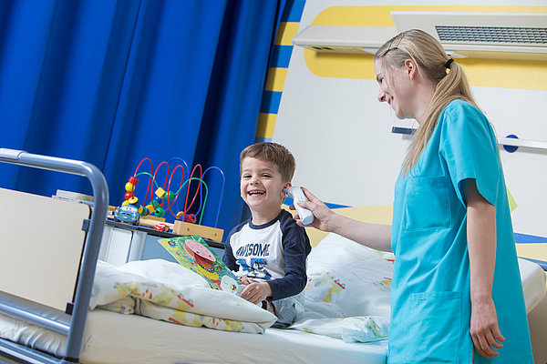 Krankenpflegerin misst Fieber bei sitzendem Kind im Krankenhauszimmer mit blauem Vorhang und Spielzeug auf dem Bett