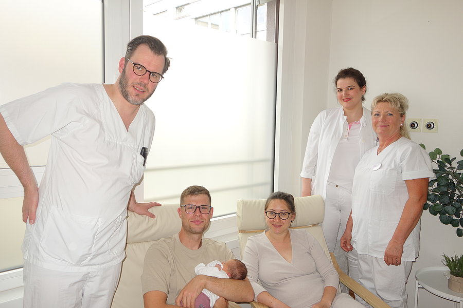 Drei in weißer Arbeitskleidung stehende Personen und ein sitzendes Paar, das ein Neugeborenes hält, in einem hellen Raum mit Fenster und Pflanze
