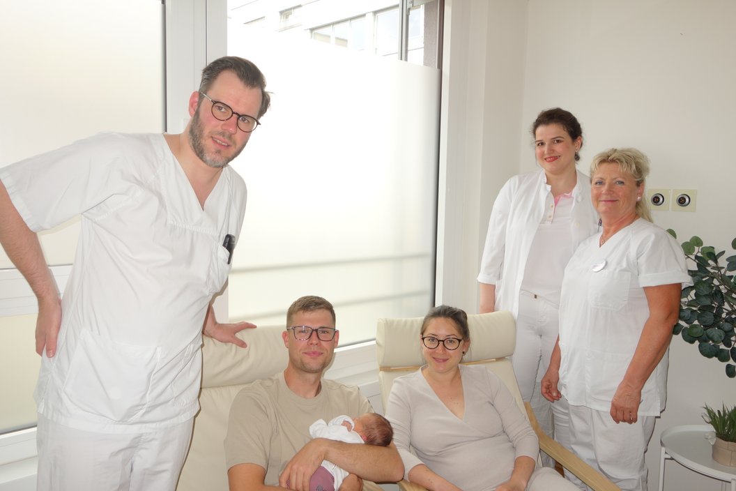 Drei in weißer Arbeitskleidung stehende Personen und ein sitzendes Paar, das ein Neugeborenes hält, in einem hellen Raum mit Fenster und Pflanze
