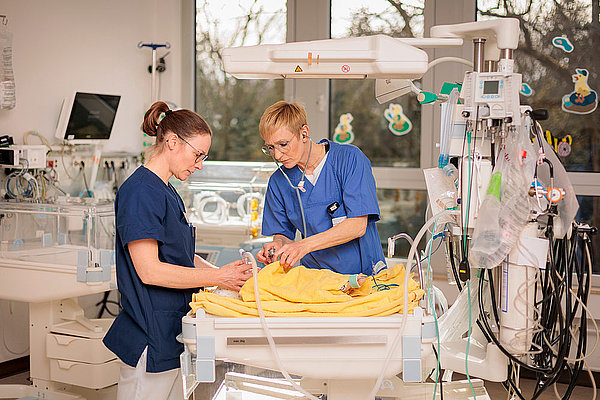 Zwei Pflegekräfte in blauer Kleidung betreuen ein Baby in einem Krankenhausbett mit medizinischen Geräten