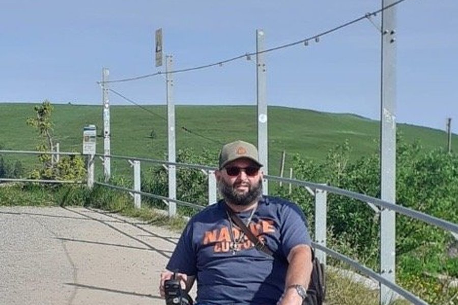 Mann mit Sonnenbrille und Basecap sitzt in einem Rollstuhl auf einem Weg mit grünem Hügel im Hintergrund
