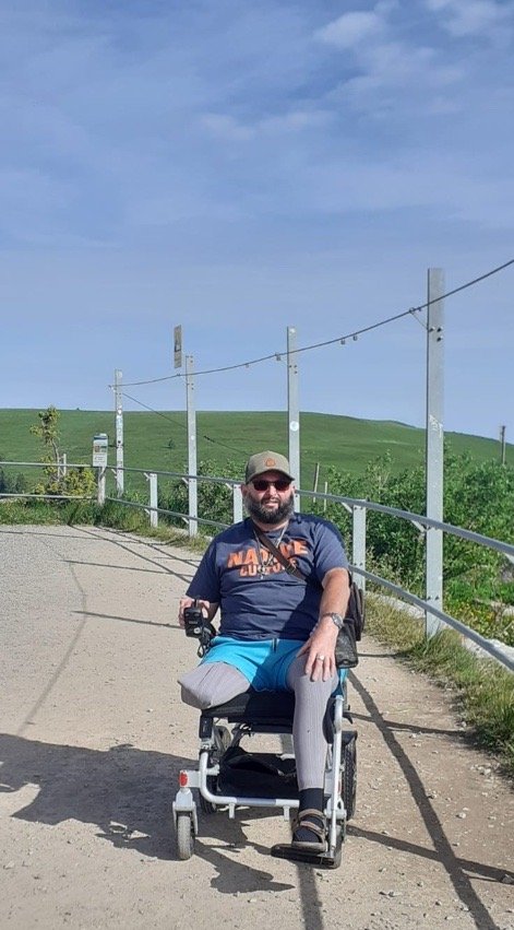 Mann mit Sonnenbrille und Basecap sitzt in einem Rollstuhl auf einem Weg mit grünem Hügel im Hintergrund