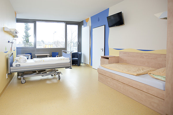 Krankenhauszimmer mit verstellbarem Patientenbett, Holzsofa, Fernseher an der Wand und großen Fenstern