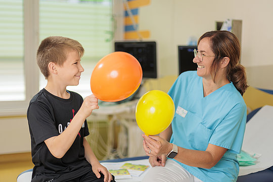 Kind und Frau in medizinischer Kleidung sitzen sich gegenüber und halten jeweils einen Luftballon