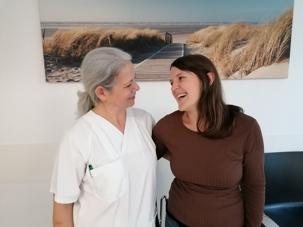 Eine Person in einem weißen Arztkittel steht neben einer Person in einem braunen Oberteil. Im Hintergrund ist ein Bild von einem Strand mit Dünen und einem Holzsteg zu sehen.