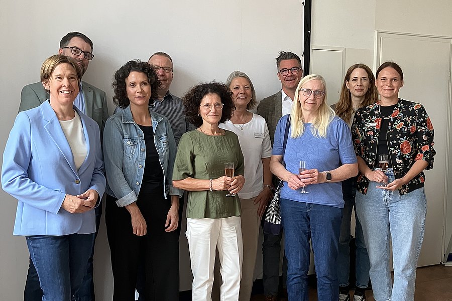 Gruppe von neun Personen steht in einem Raum vor einer weißen Wand