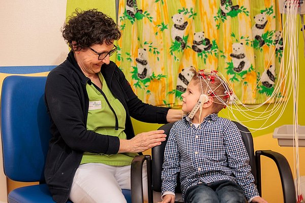Kind sitzt mit EEG-Elektroden am Kopf auf einem Stuhl, wird von medizinischer Fachkraft betreut, bunte Panda-Vorhänge im Hintergrund