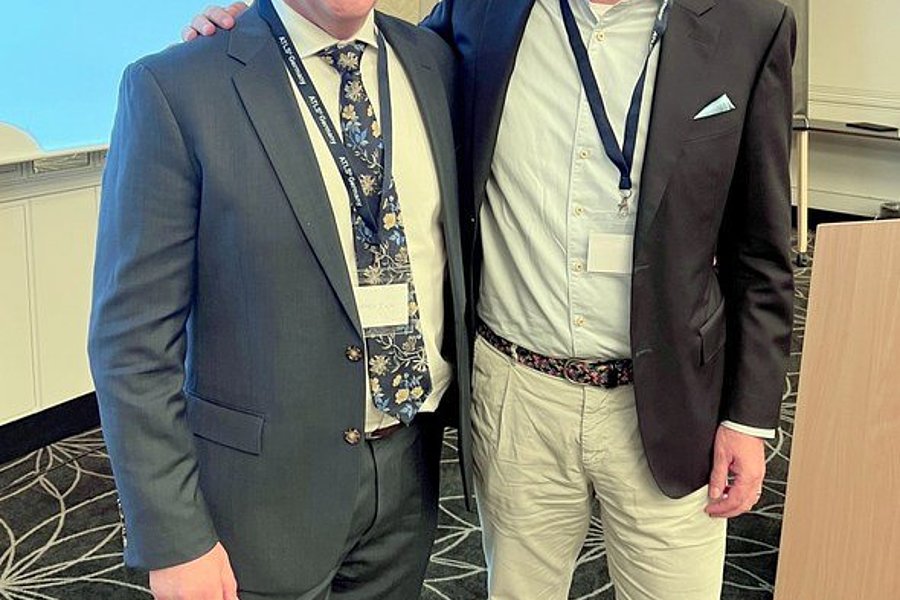 Zwei Männer stehen nebeneinander in einem Konferenzraum. Der Mann links trägt einen dunklen Anzug mit einer gemusterten Krawatte, der Mann rechts trägt eine hellere Jacke und helle Hosen.