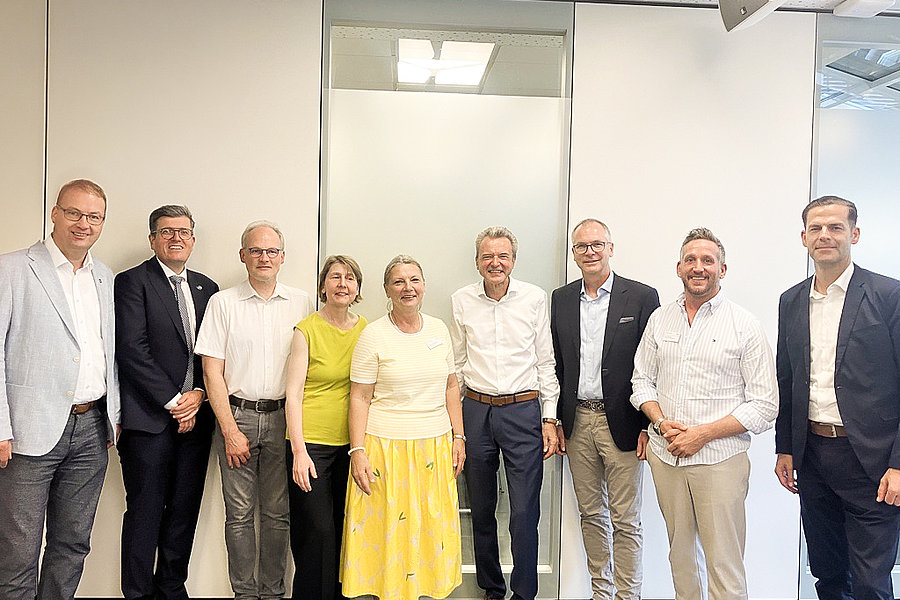 Gruppe von neun Personen steht in einem hellen Raum vor einer weißen Wand und lächelt in die Kamera