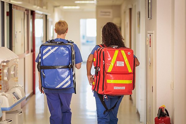 Zwei medizinische Fachkräfte in blauer Arbeitskleidung gehen einen Krankenhausflur entlang, tragen große Notfallrucksäcke in Blau und Rot