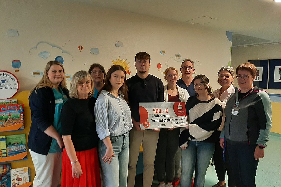 Gruppe von Personen, die vor einer Wand mit Wolken und Sonne stehen. Eine Person hält ein großes Schild mit der Aufschrift '500,- €'.