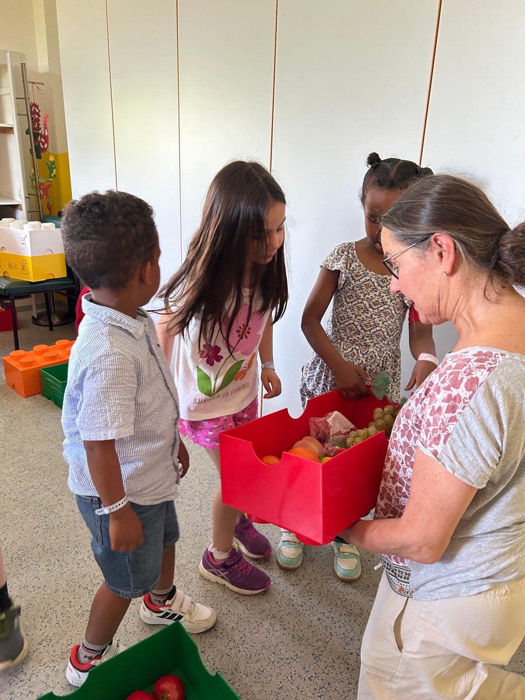 Drei Kinder und eine Frau stehen in einem Raum, die Frau hält eine rote Kiste mit Gegenständen