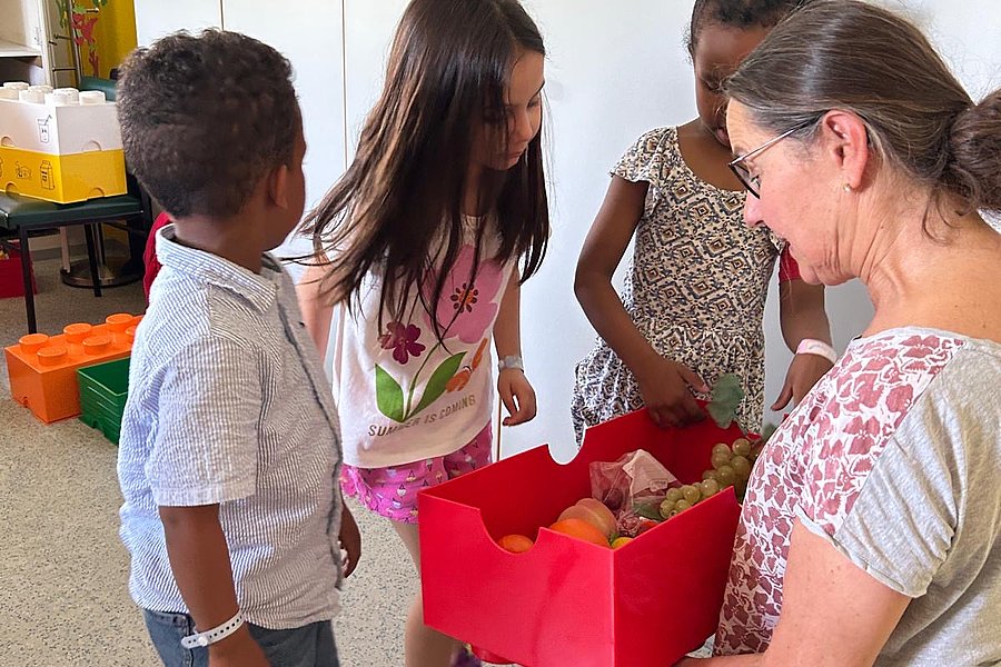 Drei Kinder und eine Frau stehen in einem Raum, die Frau hält eine rote Kiste mit Gegenständen