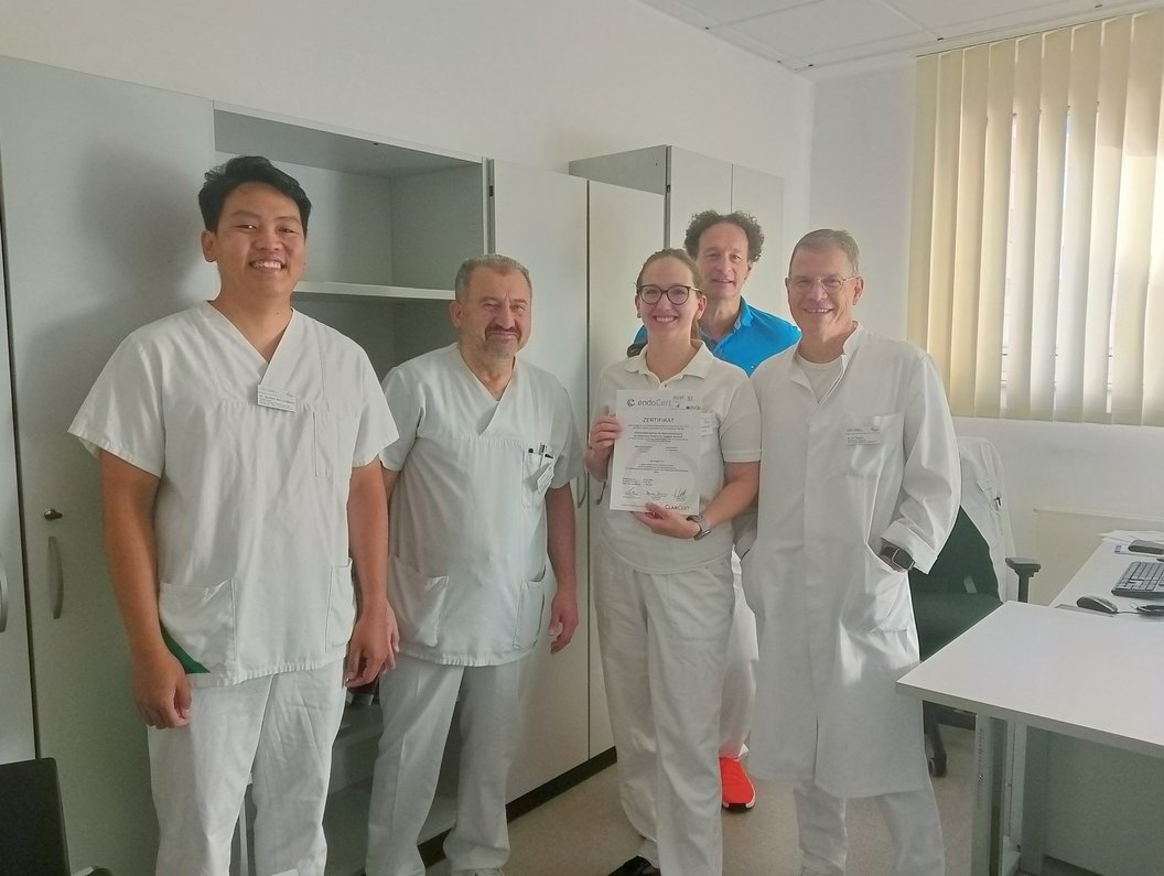 Fünf Personen in weißer Arbeitskleidung stehen in einem hellen Raum mit Schreibtisch und Fenster mit vertikalen Jalousien, eine Person hält eine Urkunde