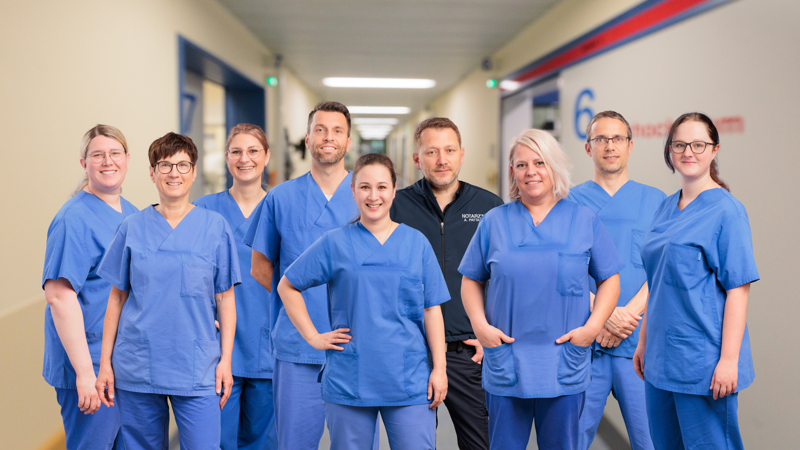 Neun medizinische Fachkräfte in blauer Arbeitskleidung stehen in einem Krankenhausflur und lächeln in die Kamera