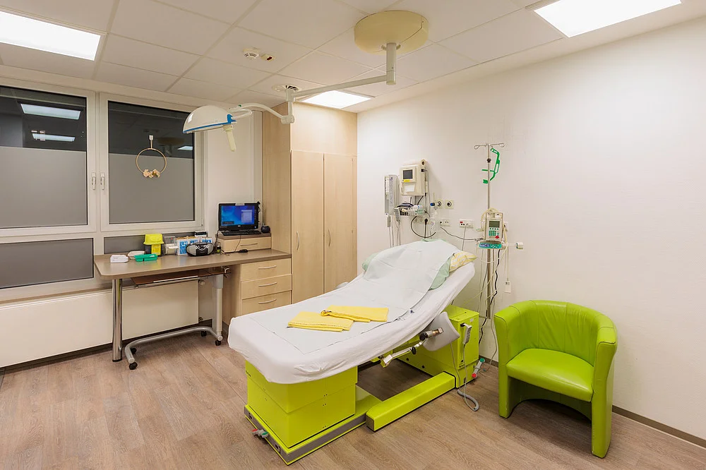 Ein Krankenhauszimmer mit einem grünen Bett, einem grünen Stuhl und einem Tisch. An der Wand sind medizinische Geräte und ein Monitor sichtbar.