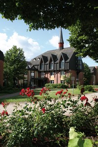 Zweistöckiges Gebäude mit spitzem Turm und Dach, umgeben von Bäumen und roten Rosen im Vordergrund.