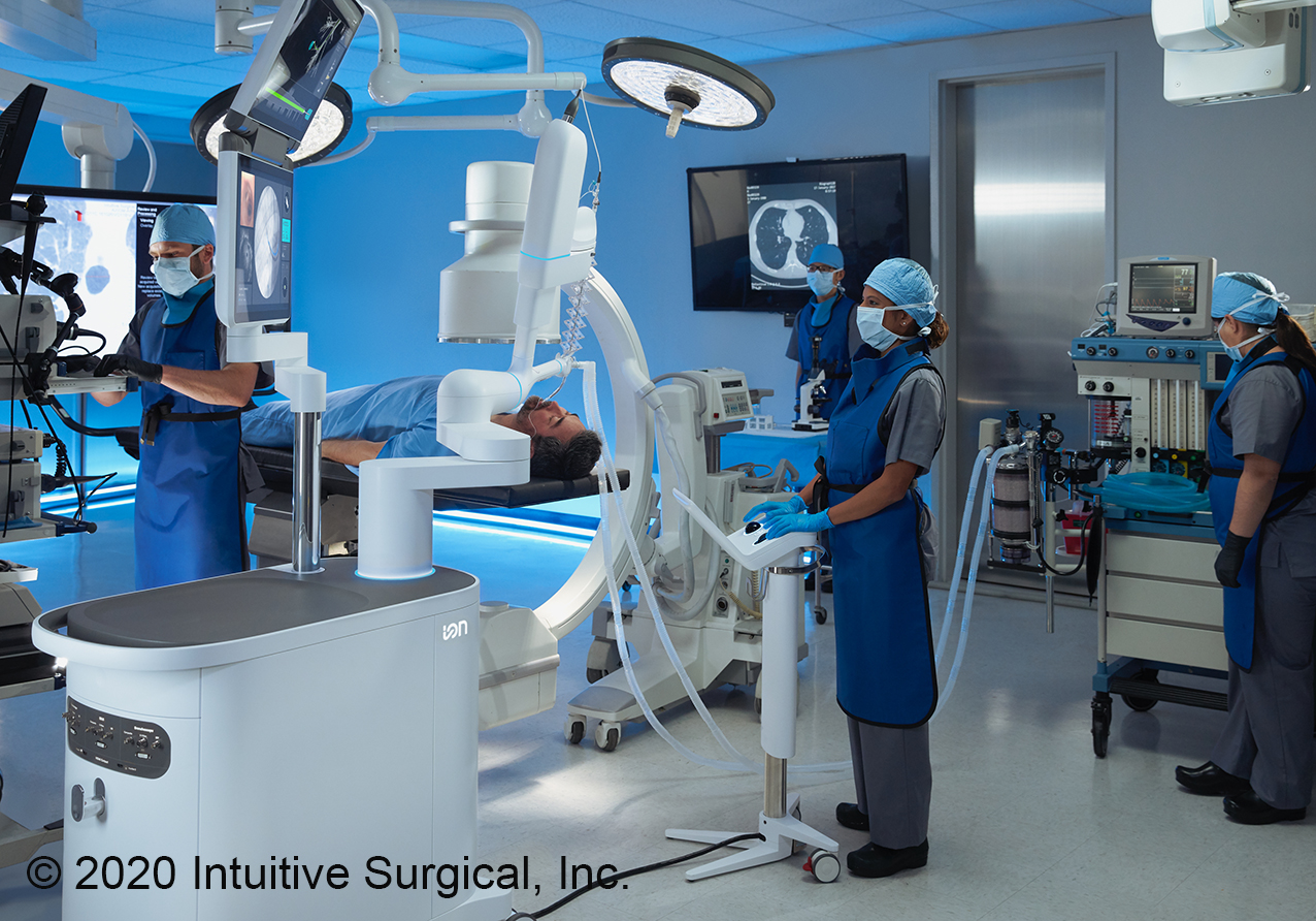 Ein medizinisches Team in blauen Kitteln und Masken arbeitet in einem Operationssaal. Ein Patient liegt auf einem Tisch, während Geräte zur Bildgebung und ein Roboterarm sichtbar sind.